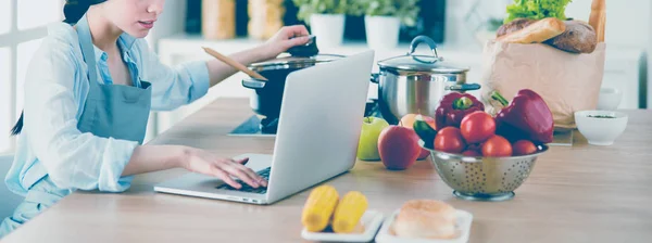 Giovane donna in cucina con computer portatile alla ricerca di ricette, sorridente. Concetto di food blogger — Foto Stock