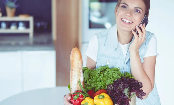 Smiling woman with mobile phone holding shopping bag in kitchen — Stock Photo, Image