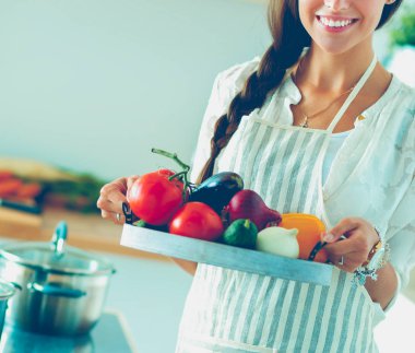 Mutfakta elinde sebzelerle gülümseyen genç bir kadın.