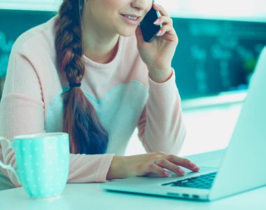 Genç kadın bir dizüstü bilgisayar kullanarak ve cep telefonuyla konuşurken mutfak masasında oturur.