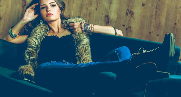 Портрет элегантной женщины, сидящей на черном диване в синих джинсах и меховом жилете — стоковое фото