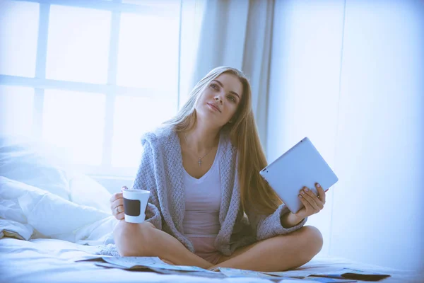 Avslappnad ung kvinna sitter på sängen med en kopp kaffe och digital tablett — Stockfoto