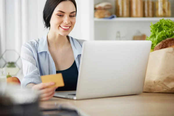 Mujer sonriente compras en línea utilizando la computadora y la tarjeta de crédito en la cocina —  Fotos de Stock