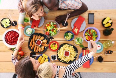 Ahşap masada otururken birlikte yemek yiyen bir grup insanın üst düzey görüntüsü. Masada yemek var. İnsanlar fast food yiyor..