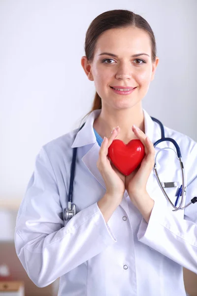 Bonito jovem sorridente médico feminino sentado na mesa e segurando o coração . — Fotografia de Stock