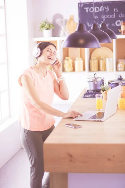 Porträtt av en glad ung kvinna lyssnar på musik med hörlurar och använda bärbar dator medan du står i köket — Stockfoto
