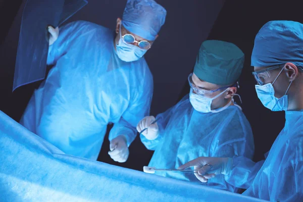 Grupo de cirurgiões no trabalho em sala de operações tonificada em azul. Equipe médica realizando operação — Fotografia de Stock