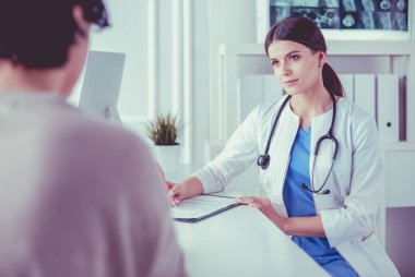 Doktor ve hasta hastane danışma odasında tıbbi sorunları tartışıyorlar. Doktor hasta formunu dolduruyor.