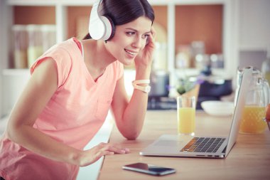 Mutfakta dururken kulaklıkla müzik dinleyen ve dizüstü bilgisayar kullanan neşeli bir genç kadının portresi