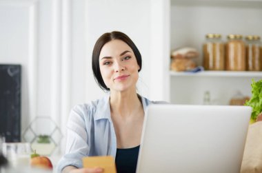 Mutfakta bilgisayar ve kredi kartı kullanan gülümseyen bir kadın.