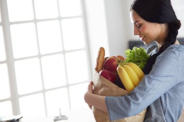 Sebzeli alışveriş poşeti taşıyan genç bir kadın. Mutfakta duruyor.