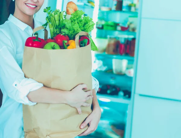 Mujer joven sosteniendo bolsa de la compra de comestibles con verduras.De pie en la cocina. Mujer en la cocina mirando a la cámara — Foto de Stock