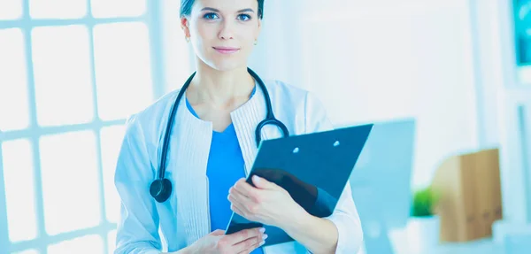 Joven doctora sonriente con estetoscopio sosteniendo una carpeta en una sala de consulta de hospitales — Foto de Stock