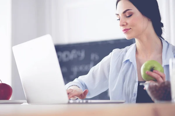 Woman with apple and laptop sitting in the kitchen