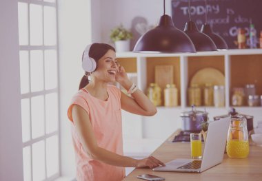 Mutfakta dururken kulaklıkla müzik dinleyen ve dizüstü bilgisayar kullanan neşeli bir genç kadının portresi