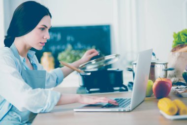 Mutfakta dizüstü bilgisayara benzeyen tariflerle gülümseyen genç bir kadın. Gıda blogcusu kavramı