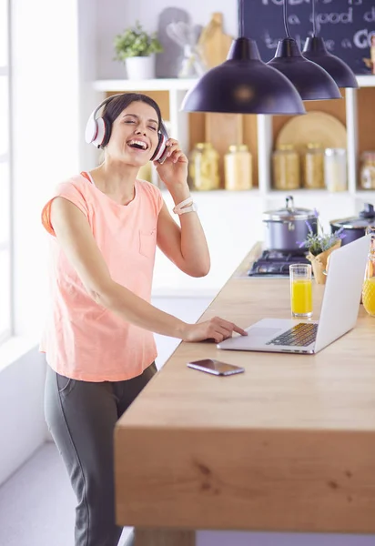 Portrait d'une jeune femme joyeuse écoutant de la musique avec des écouteurs et utilisant un ordinateur portable tout en restant debout à la cuisine — Photo