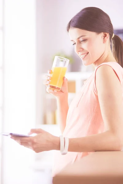 Lächelnde hübsche Frau, die beim Frühstück in der Küche auf ihr Handy schaut und ein Glas Orangensaft in der Hand hält. — Stockfoto