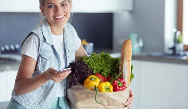 Femeie tânără care deține sac de cumpărături alimentar cu legume. Stând în bucătărie — Fotografie, imagine de stoc