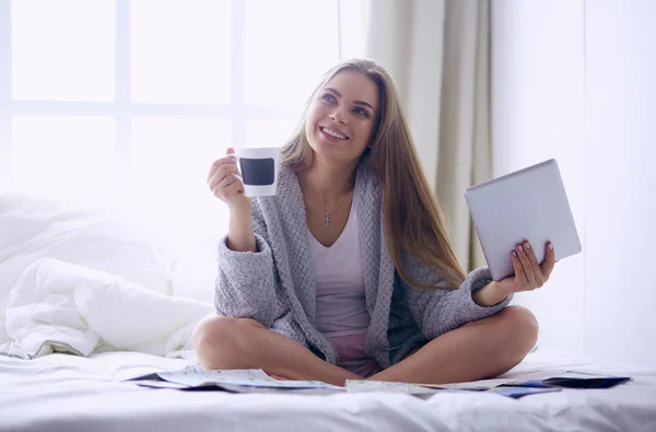 Avslappnad ung kvinna sitter på sängen med en kopp kaffe och digital tablett — Stockfoto