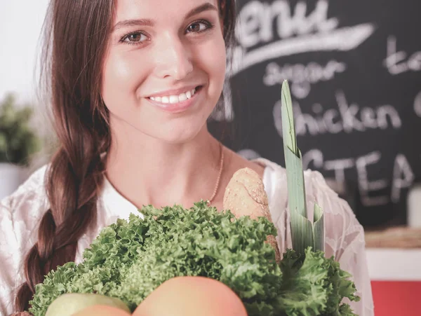Smiling young woman holding vegetables standing in kitchen. Smiling young woman