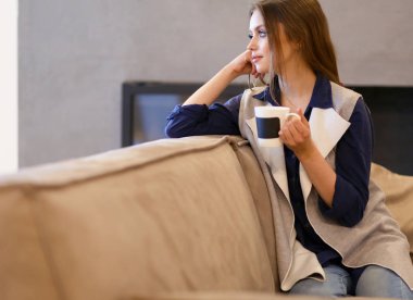 Genç bir kadın, beyaz odada rahat bir koltukta, elinde kahveyle oturuyor.