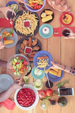 Ahşap masada otururken birlikte yemek yiyen bir grup insanın üst düzey görüntüsü. Masada yemek var. İnsanlar fast food yiyor..