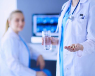 Doktorun arka planda meslektaşıyla klinikte elinde haplar ve bir bardak su tutarken görüntüsü..