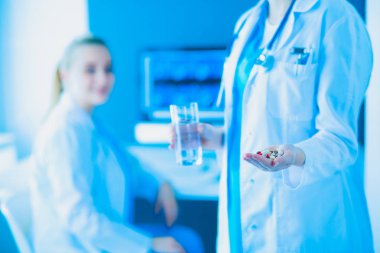 Doktorun arka planda meslektaşıyla klinikte elinde haplar ve bir bardak su tutarken görüntüsü..