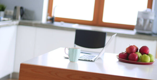 Young woman sits at the kitchen table using a laptop and talking on a cell phone.