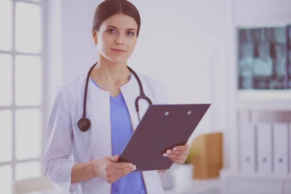 Joven doctora sonriente con estetoscopio sosteniendo una carpeta en el consultorio médico — Foto de Stock