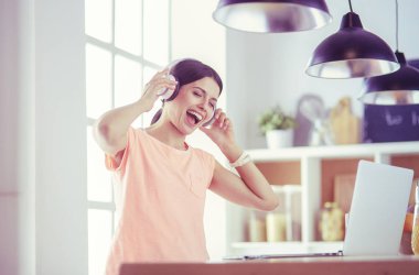 Mutfakta dururken kulaklıkla müzik dinleyen ve dizüstü bilgisayar kullanan neşeli bir genç kadının portresi