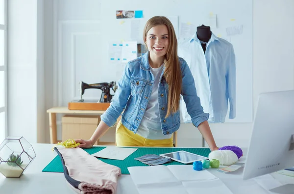 Schöne Modedesignerin steht neben Schreibtisch im Atelier — Stockfoto