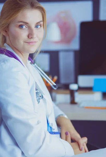 Schöne junge lächelnde Ärztin sitzt am Schreibtisch — Stockfoto