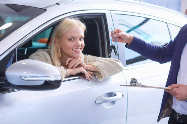 Hermosa joven mujer está recibiendo la llave y sonriendo mientras está sentado en un coche nuevo en la concesionaria — Foto de Stock