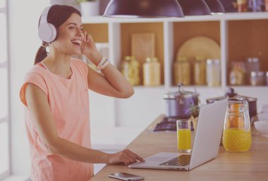 Mutfakta dururken kulaklıkla müzik dinleyen ve dizüstü bilgisayar kullanan neşeli bir genç kadının portresi