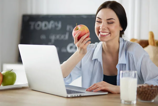 Kobieta z jabłkiem i laptopem siedzi w kuchni — Zdjęcie stockowe