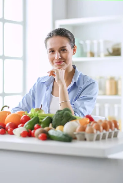 Young and cute woman sitting at the table full of fruits and vegetables in the wooden interior. — Stock Photo, Image