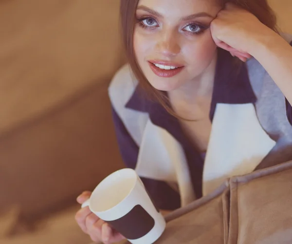 편안 한 소파에 앉아 커피를 손에 들고 하얀 방에 앉아 있는 젊은 여자 — 스톡 사진
