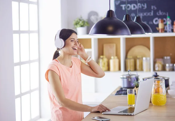 Retrato de una joven alegre escuchando música con auriculares y usando un ordenador portátil mientras está de pie en la cocina — Foto de Stock