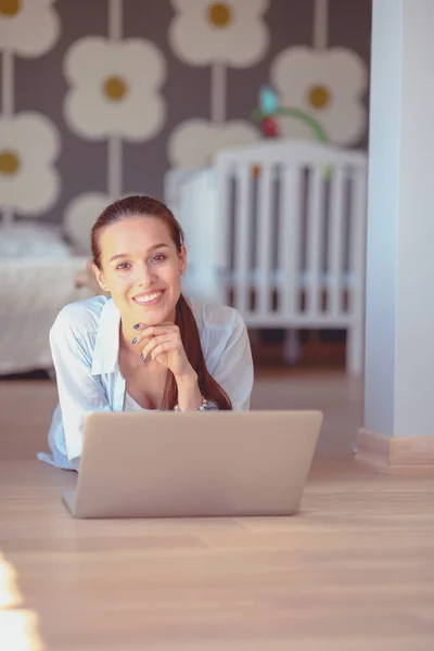 Ung kvinna som sitter på golvet nära barnens babysäng med laptop. Ung mamma — Stockfoto