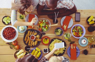 Ahşap masada otururken birlikte yemek yiyen bir grup insanın üst düzey görüntüsü. Masada yemek var. İnsanlar fast food yiyor..