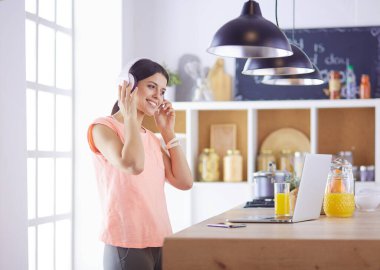 Mutfakta dururken kulaklıkla müzik dinleyen ve dizüstü bilgisayar kullanan neşeli bir genç kadının portresi