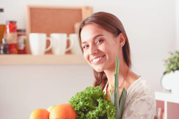 Mujer joven sonriente sosteniendo verduras de pie en la cocina. Jovencita sonriente —  Fotos de Stock