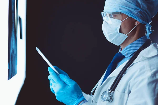 Arzt im Krankenhaus sitzt am Schreibtisch und betrachtet Röntgenbilder auf Tablet vor weißem Hintergrund mit Röntgenaufnahmen — Stockfoto
