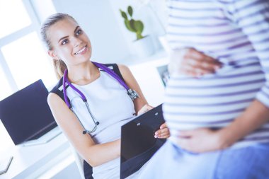 Steteskoplu ve tabletli genç kadın doktor hastanede hamile bir kadınla konuşuyor..