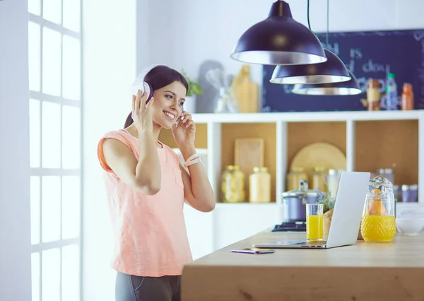 Portret van een vrolijke jonge vrouw die naar muziek luistert met een koptelefoon en met laptop computer terwijl hij in de keuken staat — Stockfoto