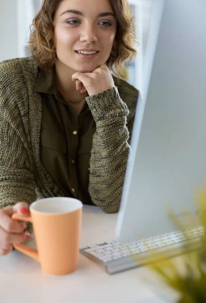 Attraktiv leende kvinna sitter vid skrivbordet, håller en kopp kaffe, hon är avkopplande och tittar bort — Stockfoto