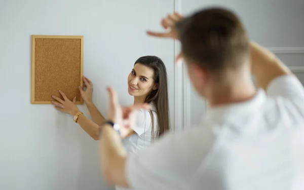 Vrouw helpt haar partner om schilderij te hangen aan de muur van hun nieuwe huis, ze is framing de foto met haar vingers — Stockfoto