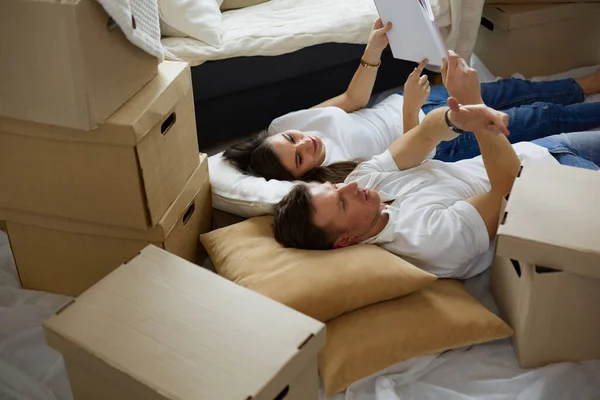 Linda pareja desempacando cajas de cartón en su nuevo hogar, tumbados en el suelo y mirando un álbum familiar —  Fotos de Stock